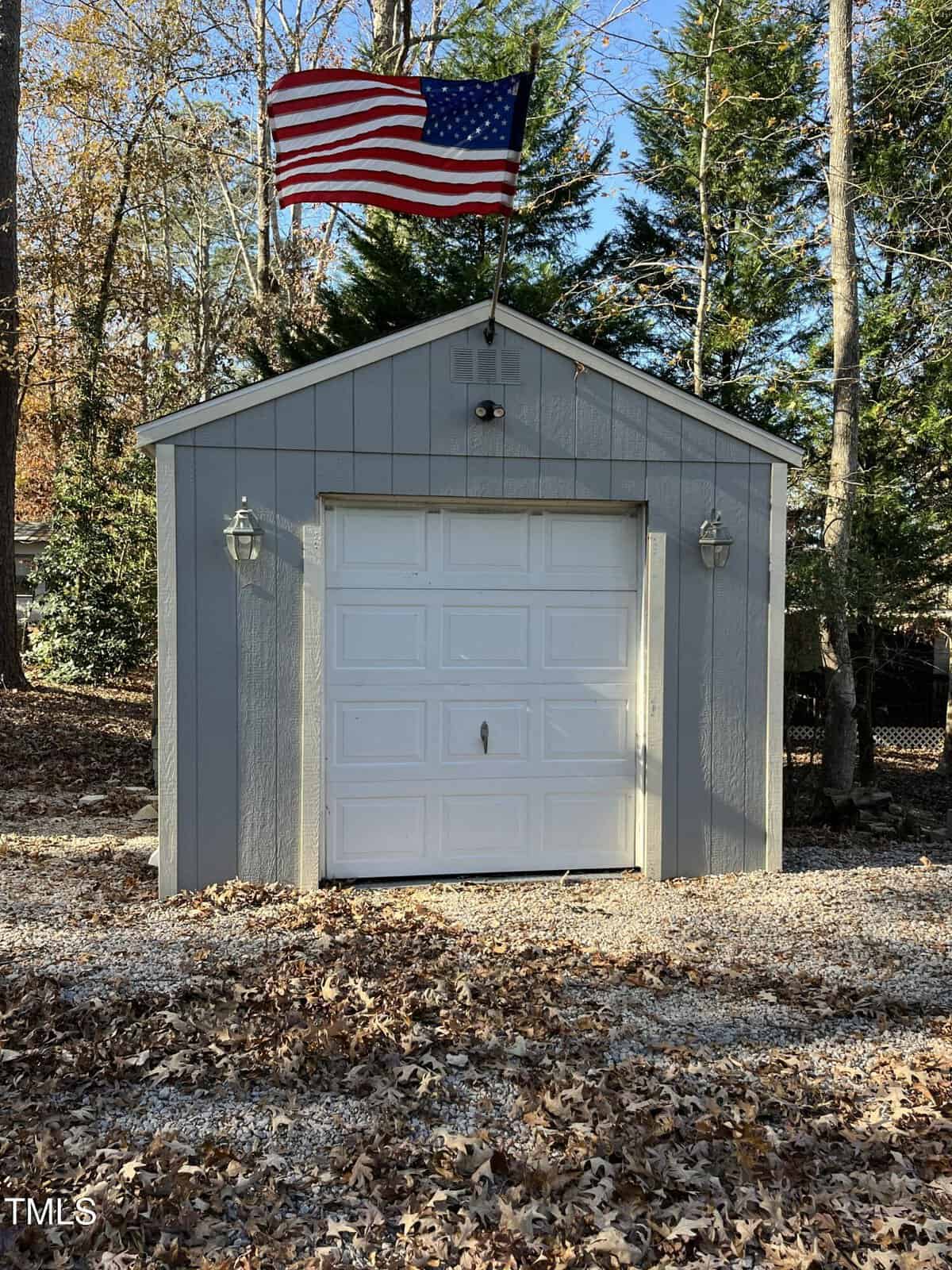 A small gray shed with a white garage door stands on a gravel lot covered with fallen leaves. An American flag is mounted on top. Two outdoor lights flank the door. Tall trees with autumn foliage are in the background.