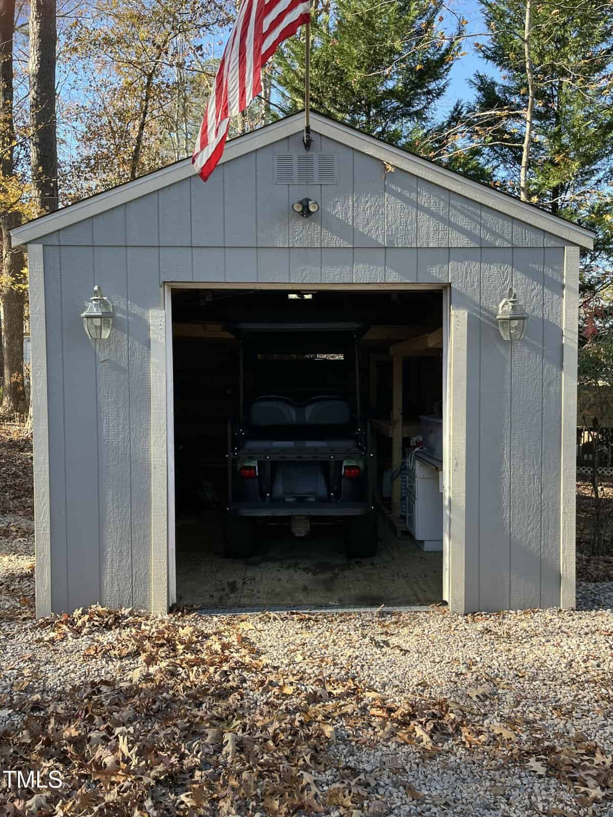 A gray shed with an open door revealing a black golf cart inside. An American flag flies above the shed, which is surrounded by trees and fallen leaves on the ground. Two lantern-style lights are mounted by the entrance.