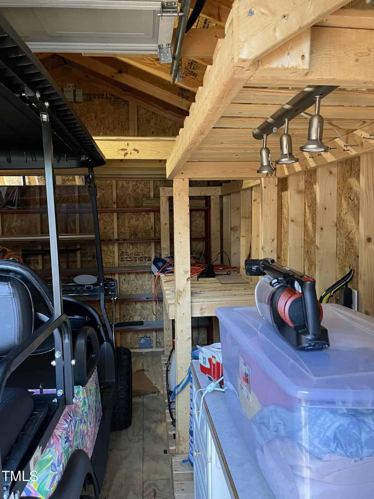A wooden shed interior with shelves, plastic storage bins, a parked golf cart, tools, and a bench with overhead lights, all organized within the unfinished wood structure.
