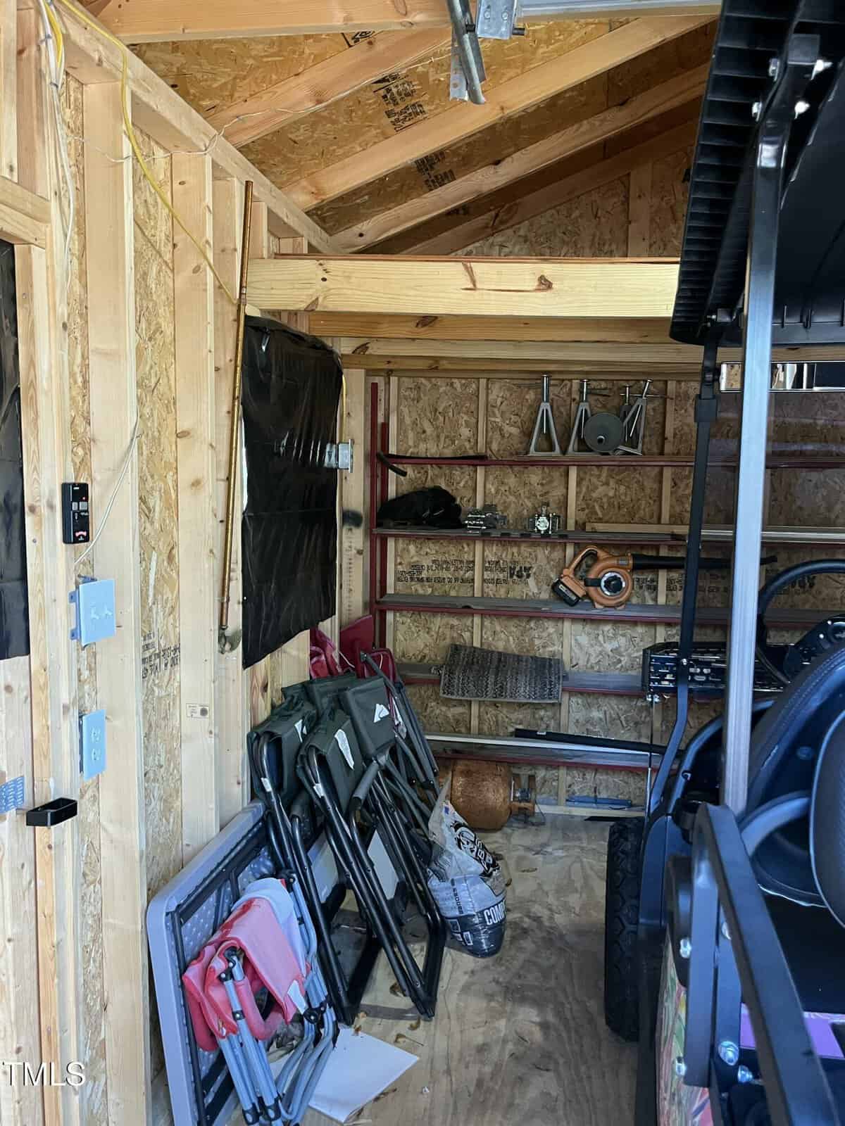 Interior of a wooden shed with folding chairs stacked on the left, shelves with various tools and items at the back, and part of a utility vehicle visible on the right side.