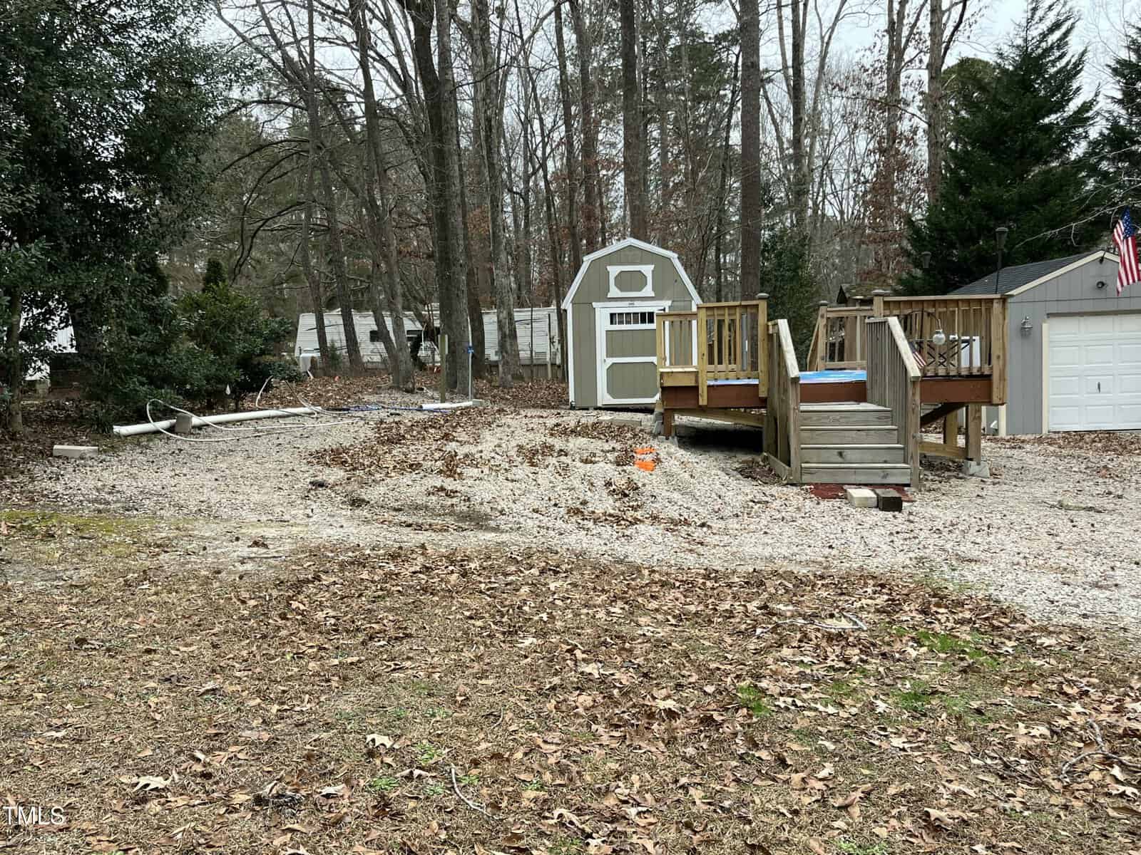 A backyard with a raised wooden deck, a small shed, a white garage, and scattered gravel and leaves. There are trees without leaves in the background and an American flag attached to the garage.