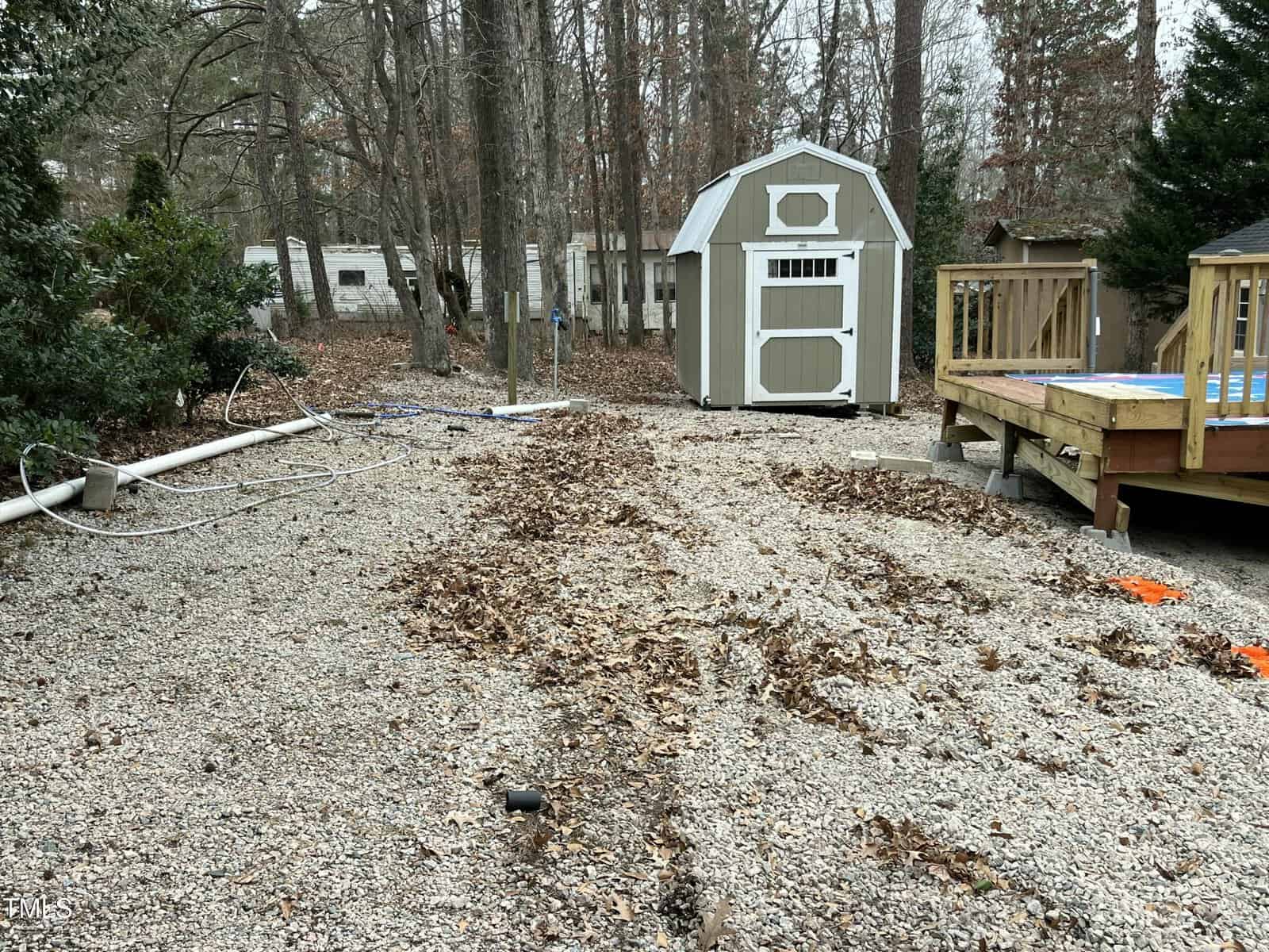 A backyard with scattered leaves and gravel, a small shed in the background, and a wooden deck with a blue-covered pool on the right. Exposed white pipes and some tools are visible on the ground.