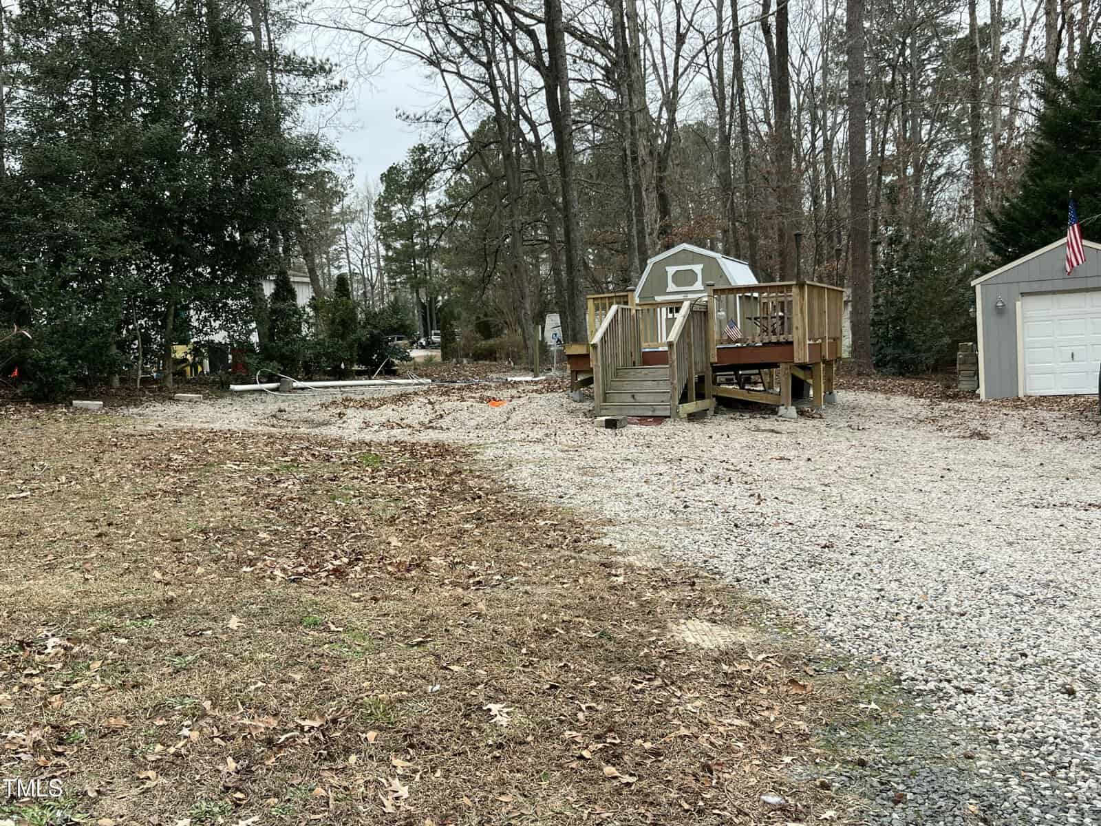 A gravel driveway leads to a small raised wooden deck beside a shed and a garage. Leafless trees and scattered leaves cover the ground. An American flag hangs on the garage. Overcast sky.