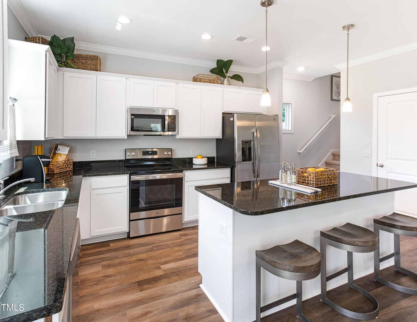 A modern kitchen with white cabinets, stainless steel appliances, black granite countertops, a kitchen island with three wooden stools, and pendant lighting. Baskets and plants decorate the upper cabinets.
