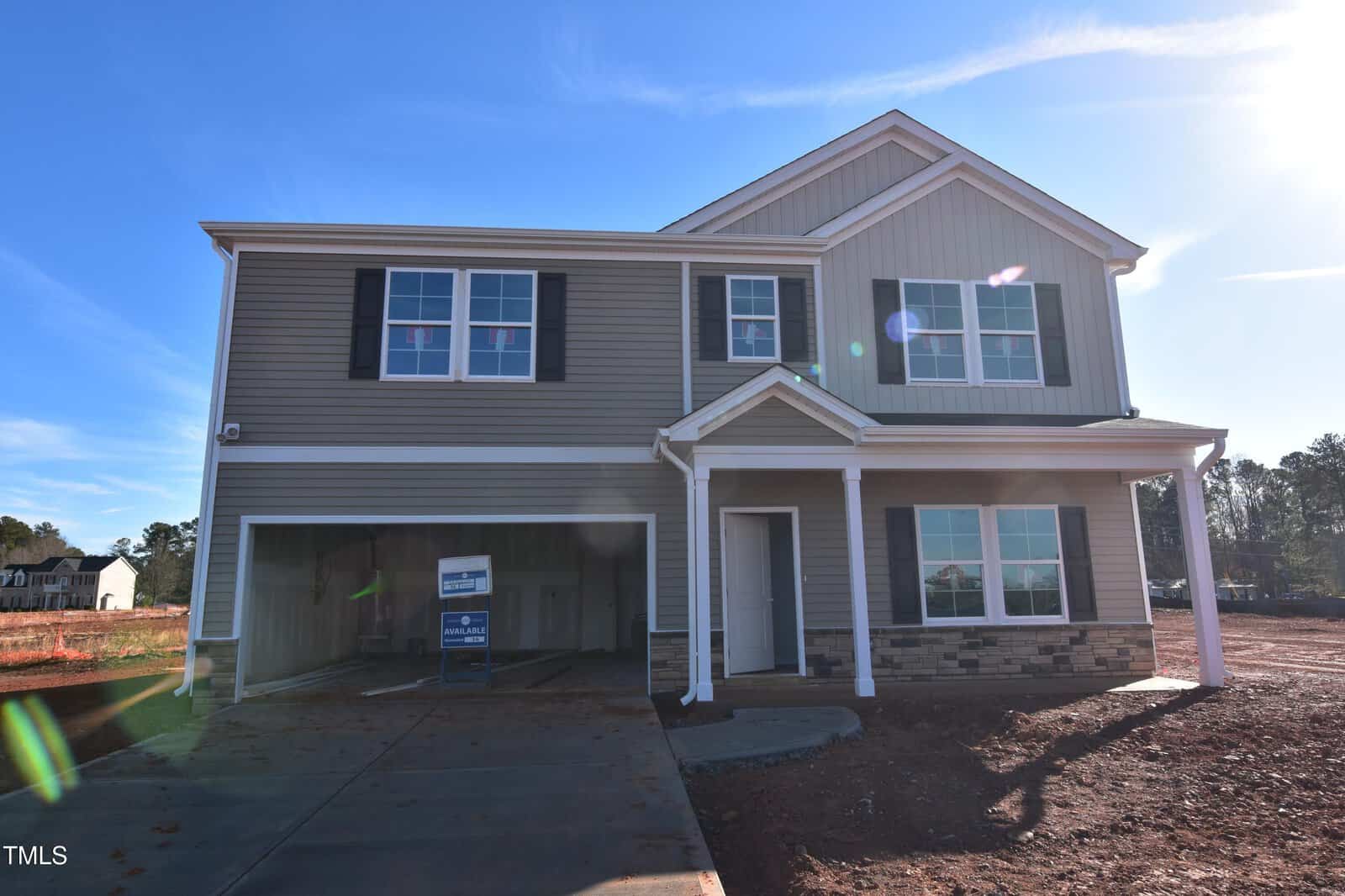 A newly constructed two-story house with beige siding, white trim, black shutters, and a covered front porch. The garage door is open, and a real estate sign is visible inside. The yard is unfinished with exposed soil.