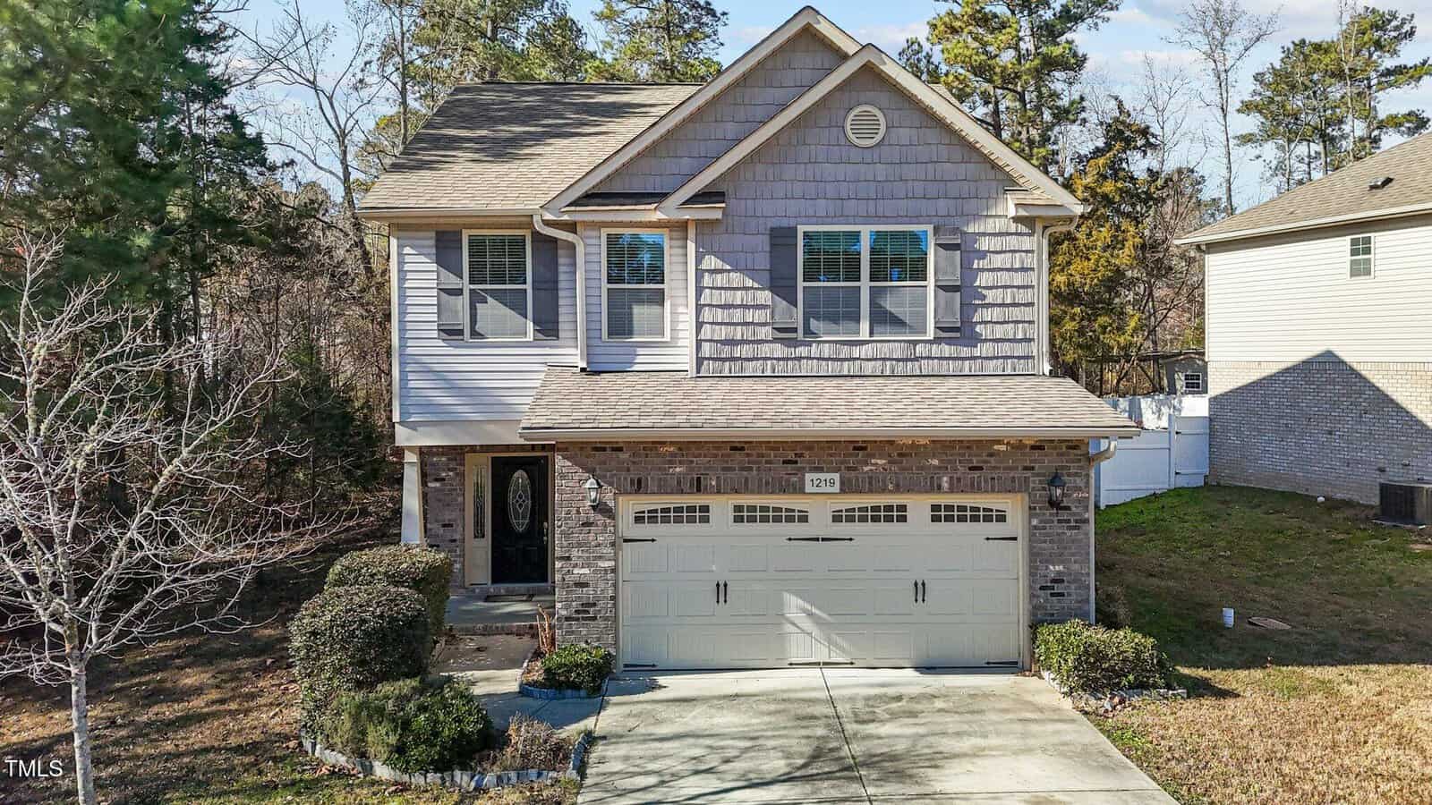 Nestled at 1219 Shining Water Lane, this charming two-story house in Creedmoor features a brick and shingle exterior, a double garage, and a concrete driveway. The landscaped yard boasts bushes and a small tree, perfectly complementing the suburban backdrop with trees in the distance.