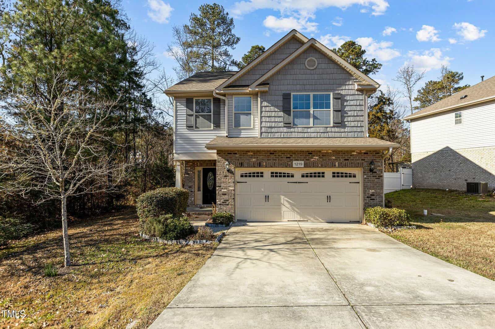 A two-story house with a light gray exterior, brick accents, and a double garage. The driveway is concrete, and the yard features grass and trees. The sky is clear with a few clouds.