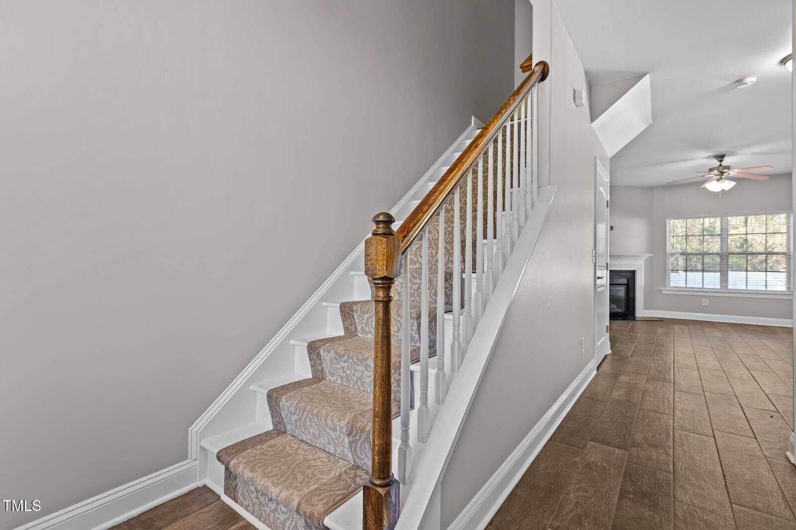 A staircase with a wooden handrail and carpeted steps leads to the second floor. The adjacent room features wood flooring, a ceiling fan, and a fireplace with large windows allowing natural light to enter. The walls are painted a light neutral color.