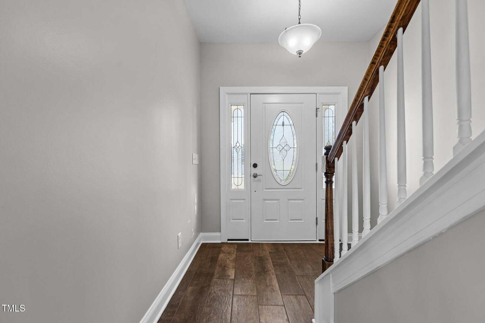 A hallway featuring light green walls, a wooden stair rail, and a dark wood floor. The white front door has an oval glass panel with decorative patterns, and a hanging pendant light illuminates the area.