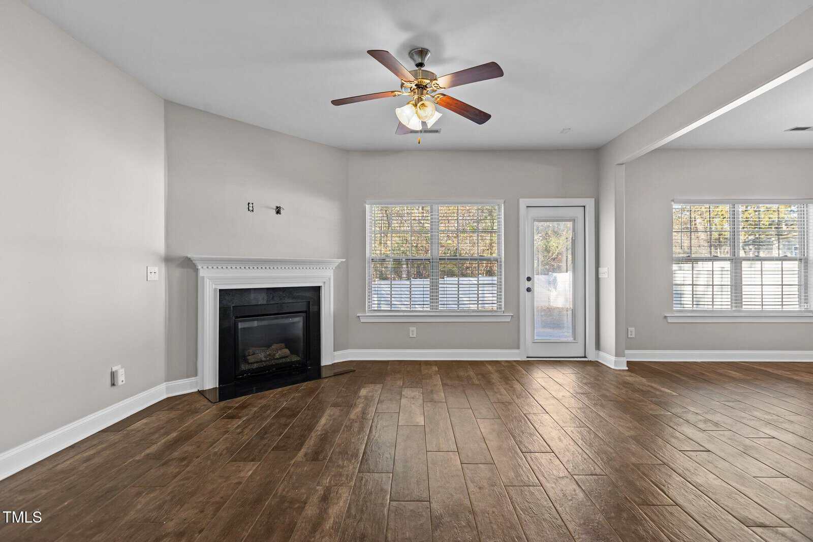 A spacious living room with light gray walls, a wooden floor, and a ceiling fan. A fireplace is on the left. Two large windows and a glass door on the right side allow natural light to enter the room.