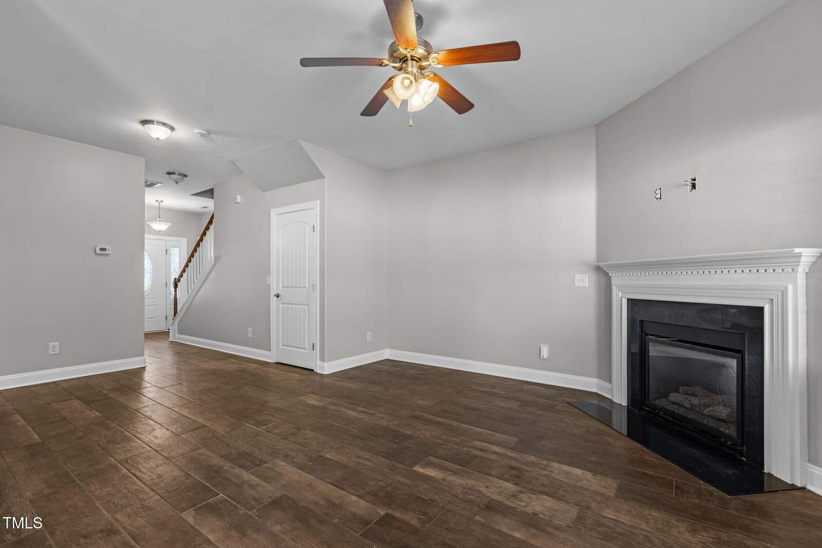A spacious living room with wooden flooring, a ceiling fan with lights, and an empty wall mount for a television above a white-trimmed fireplace. A white door leads to a hallway with stairs visible in the background.