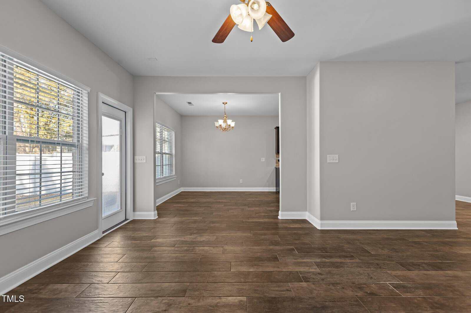 An empty room with light gray walls and wooden flooring. A ceiling fan with wooden blades hangs above. Sunlight comes in through large windows and a glass door. A chandelier is visible in an adjoining room.
