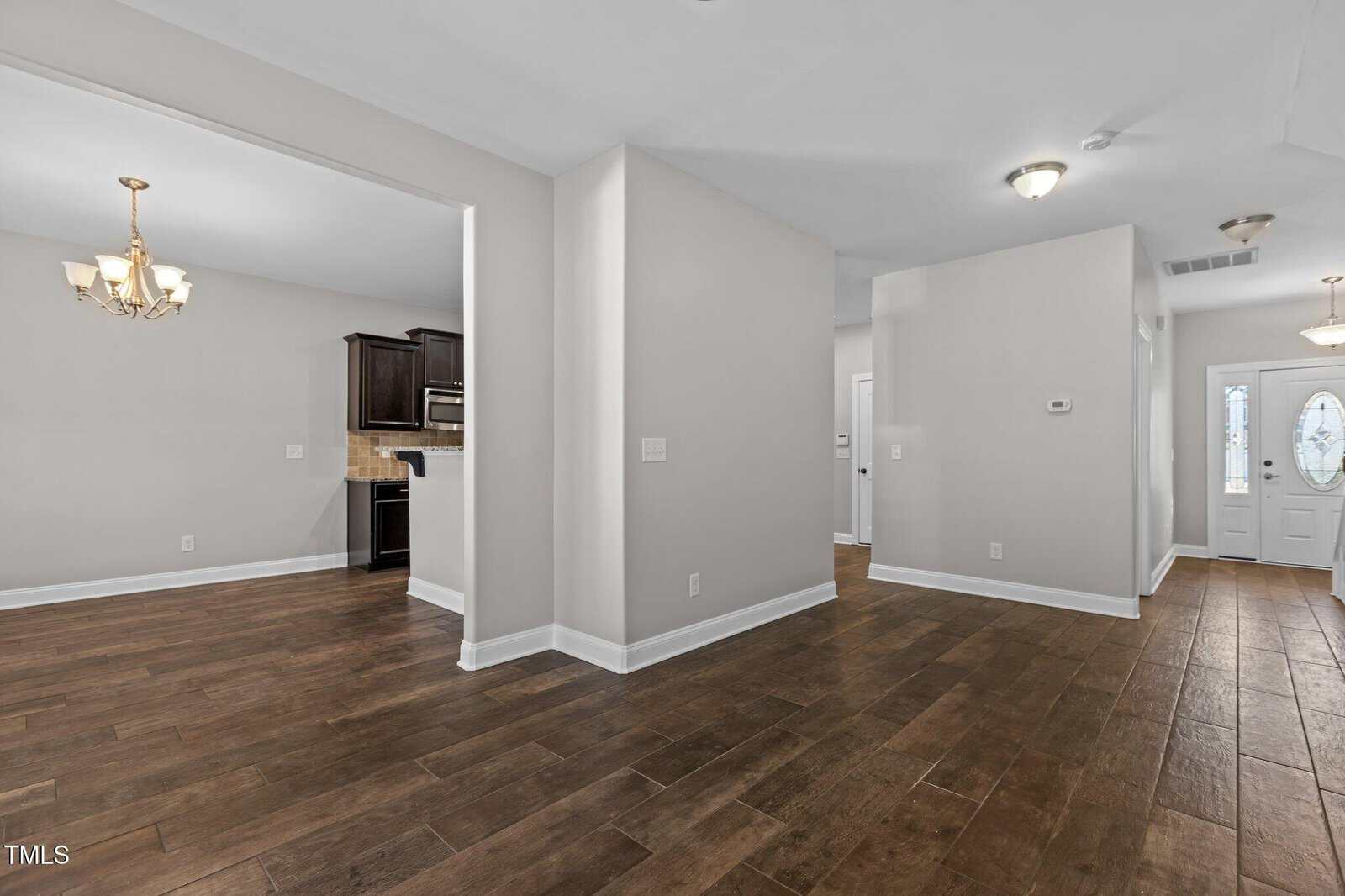 An empty room with dark wood flooring and light gray walls. A doorway leads to a kitchen area with dark cabinets. A chandelier hangs in the adjoining room. A white front door with glass panels is visible.