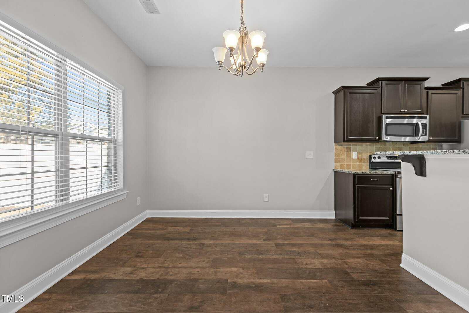 A kitchen and dining area with wooden flooring and light gray walls. The kitchen features dark brown cabinets, a stove, microwave, and a tile backsplash. A window with blinds is on the left, and a ceiling light fixture hangs above.