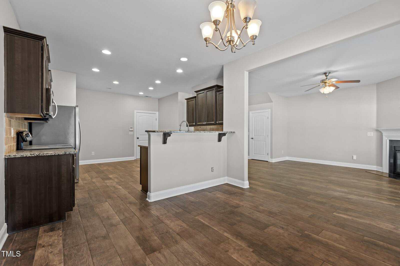 Spacious open-plan kitchen and living area with wooden floors. The kitchen features dark cabinetry, stainless steel appliances, and a central island with a granite countertop. A ceiling fan and a fireplace are visible in the adjoining living space.