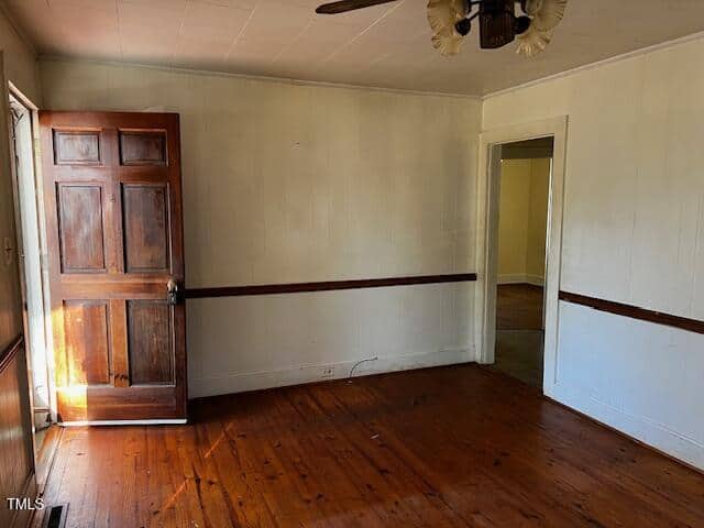 A room with wooden flooring, a dark wooden door on the left partially ajar, and a ceiling fan with a light fixture. The white walls have a dark wooden chair rail, and there's a doorway leading to another room on the right. Natural light streams in.