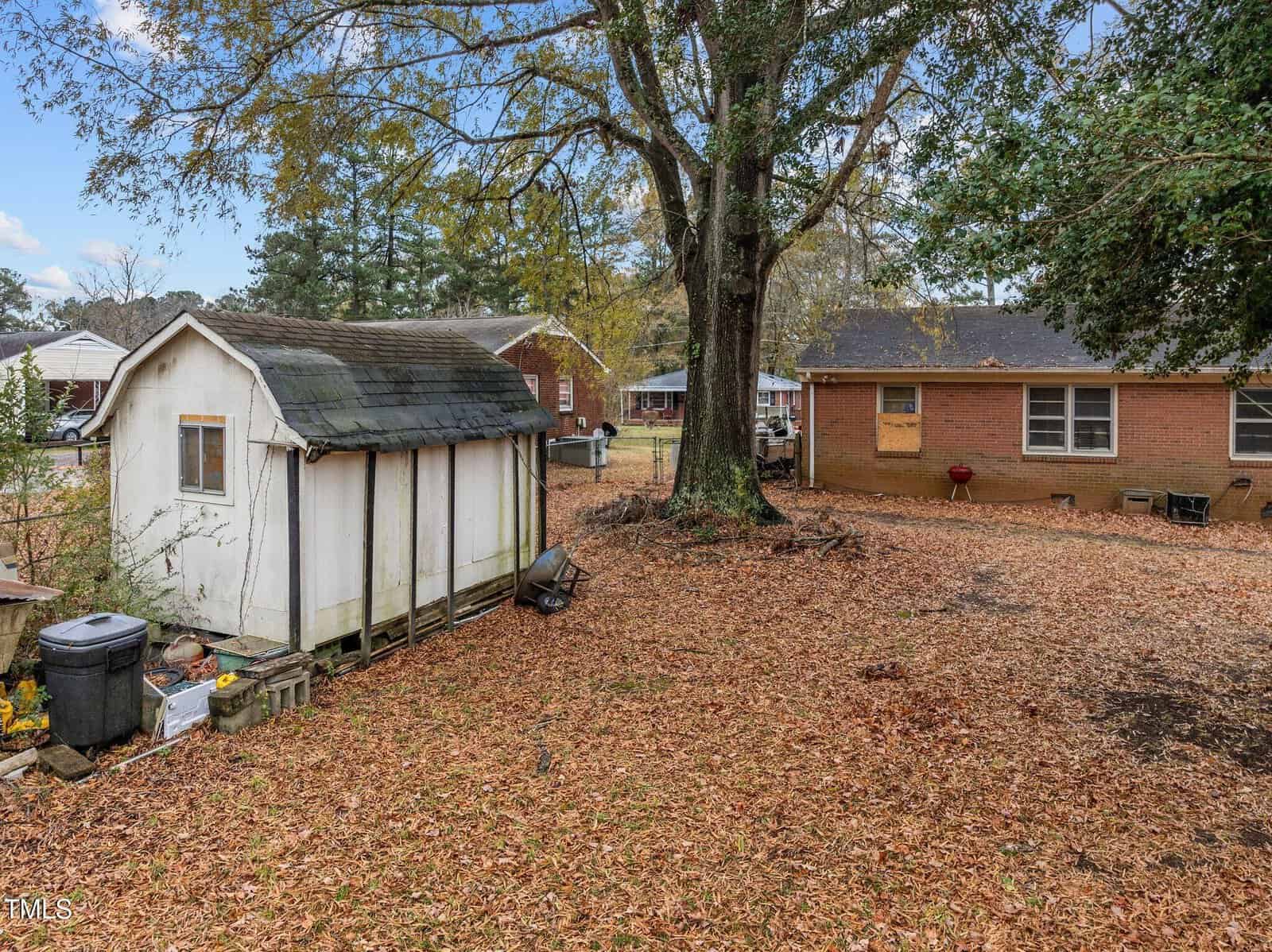 A backyard with a small white shed on the left, a large tree in the center, and a brick house in the background. The ground is covered with fallen leaves, and some gardening tools and bins are visible near the shed.
