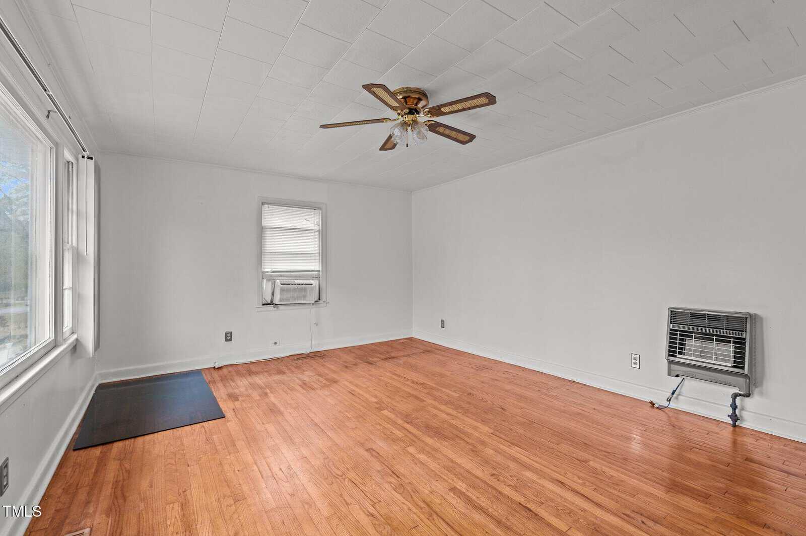 A spacious, empty room with white walls and a wooden floor. There's a ceiling fan, a window air conditioning unit, and a wall-mounted heater. Large windows on the left allow natural light to fill the room.
