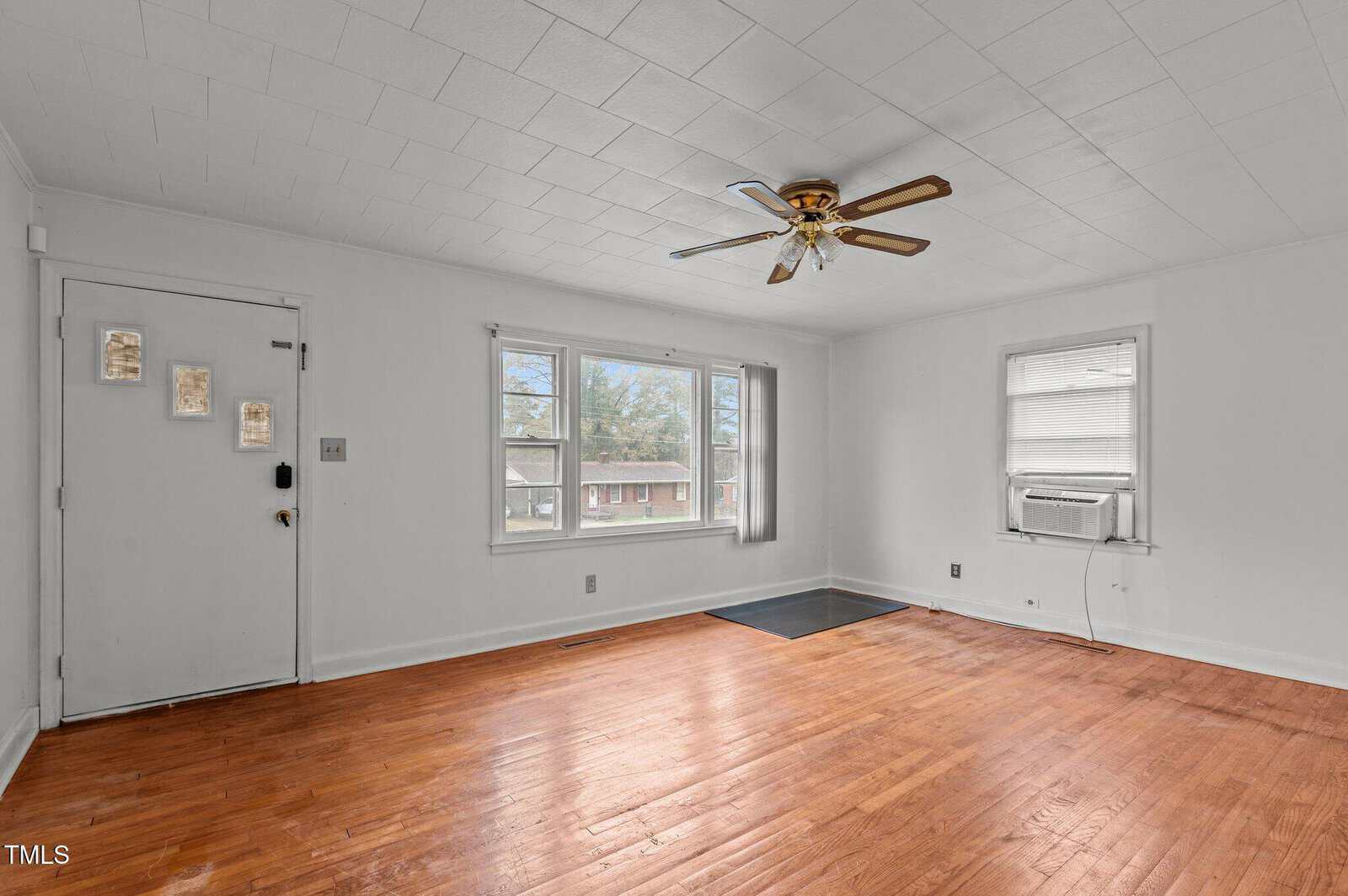 Empty room with wooden floors, a ceiling fan, and white walls. Large windows let in natural light. An air conditioning unit is installed in one window. The front door has decorative glass panels.