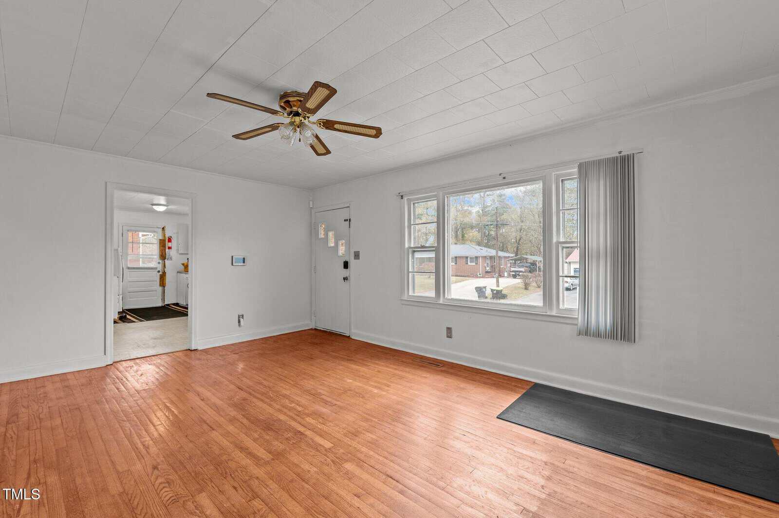 A bright, empty living room with wooden floors and a ceiling fan. Large windows on the right let in natural light. An open doorway leads to another room, hinting at a kitchen area with visible windows.