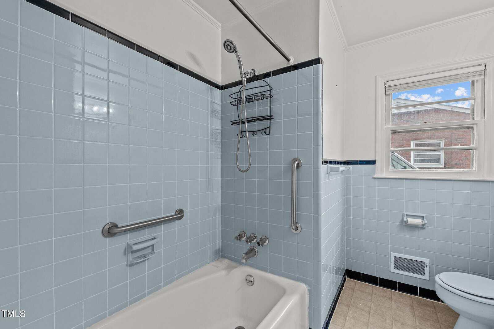 A bathroom featuring light blue tiled walls, a white bathtub with a showerhead, and a window with a view of a building outside. The bathtub includes a grab bar and built-in soap holder. The floor is tiled in beige.
