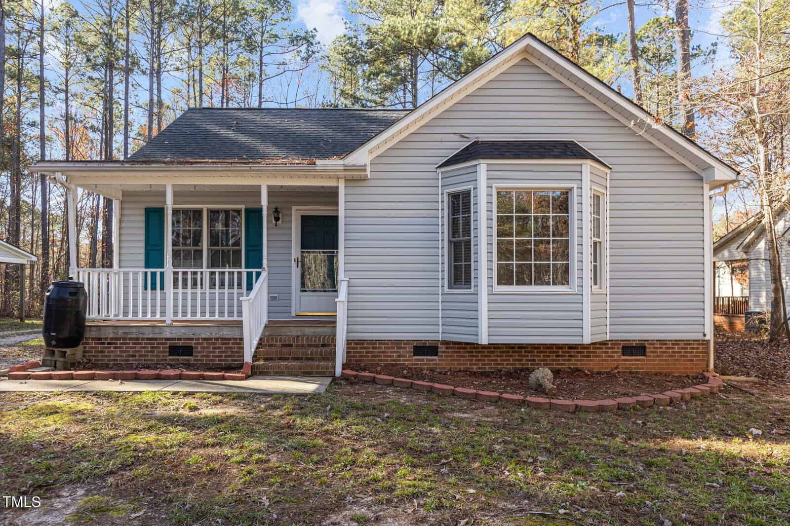 A single-story house with light gray siding, a brick foundation, a small covered front porch with white railings, blue shutters, and a bay window. Tall pine trees are visible in the background.