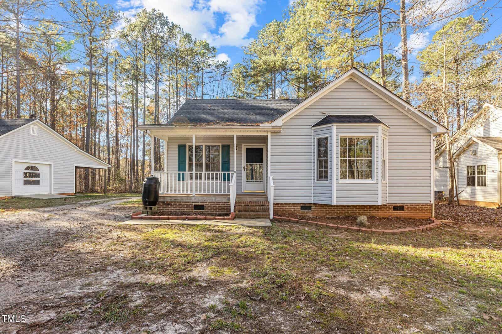 A small, single-story house with light gray siding, a covered front porch, and a bay window. The house sits on a dirt and grass lot, surrounded by tall pine trees under a partly cloudy sky.
