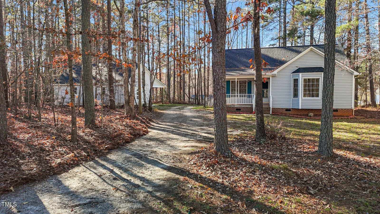 A white single-story house with a covered porch sits among tall trees. A curved gravel driveway leads to a detached garage in the background. Fallen leaves cover the ground under the trees.