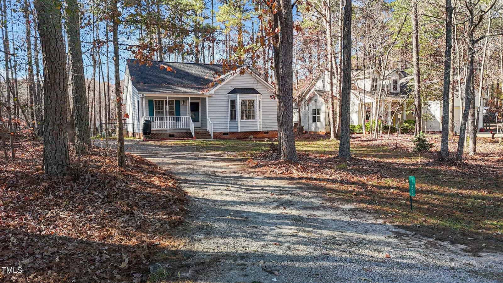 A small white house with a front porch sits among tall trees. A curved gravel driveway leads to the house, and fallen leaves are scattered on the ground. Another house is visible in the background.