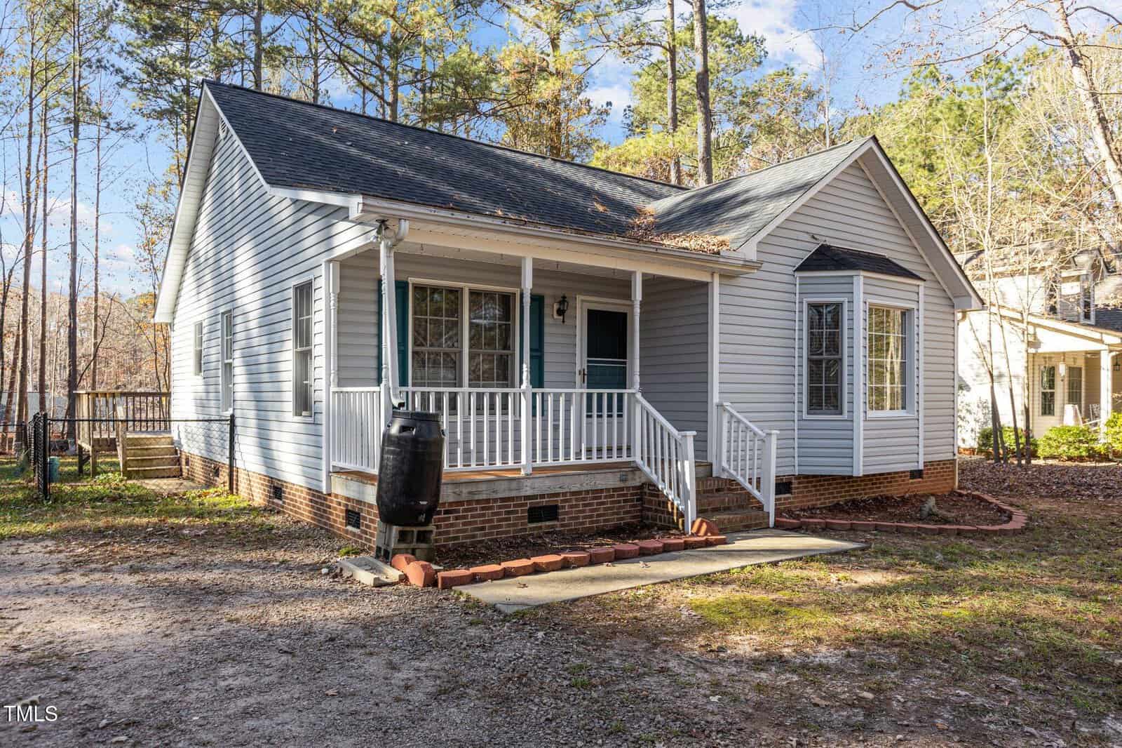 A single-story house with light gray siding, white trim, and a covered front porch with steps. A rain barrel is positioned near the porch, and the house is surrounded by trees and a gravel driveway.