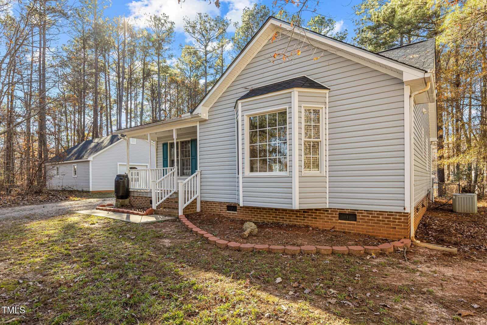 Single-story house with light gray siding, white trim, a brick foundation, front porch, bay window, and surrounded by tall trees in a wooded yard.