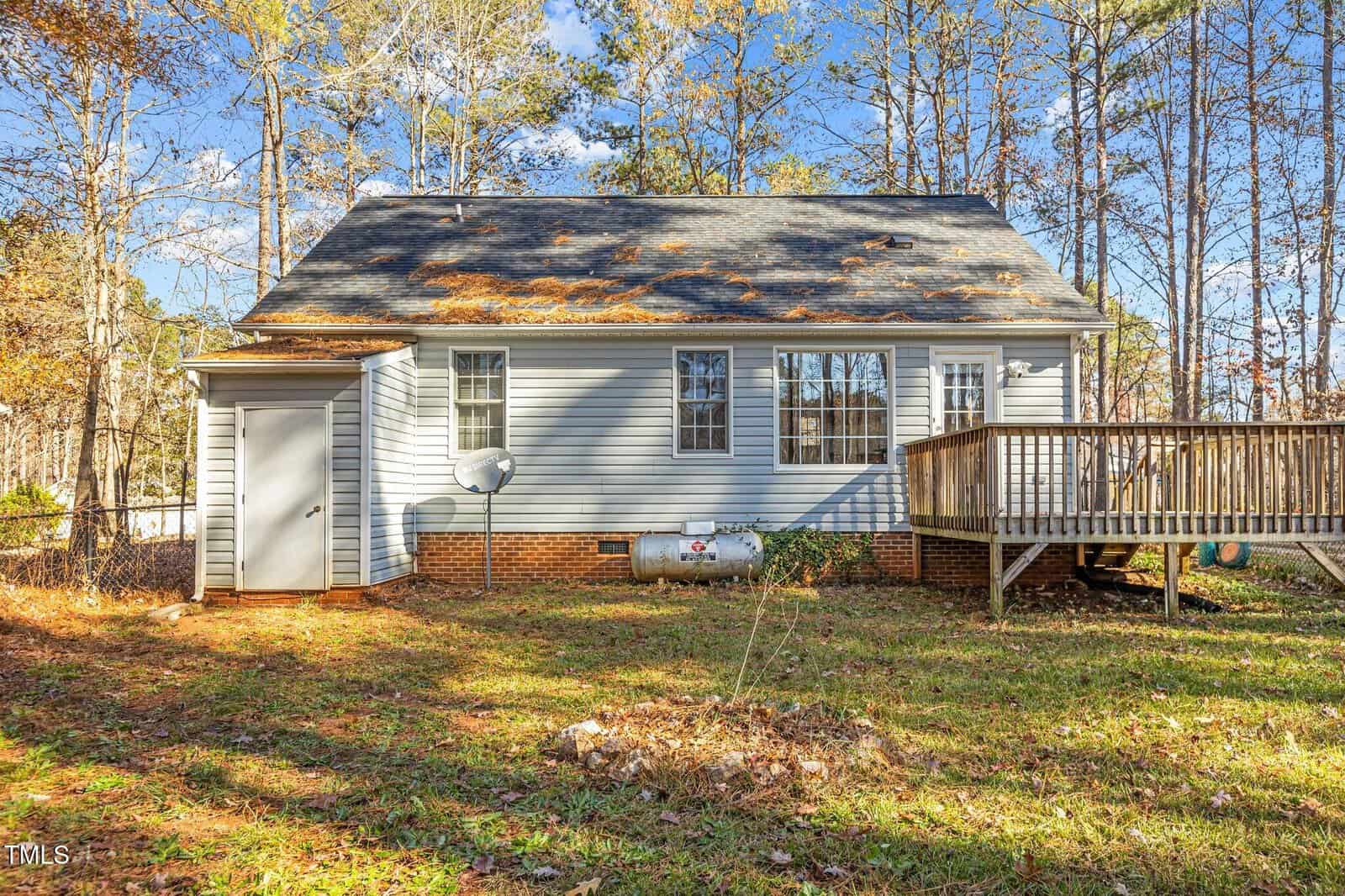 Small gray house with a sloped roof, back deck, propane tank, and satellite dish, surrounded by trees and fallen leaves on a sunny autumn day.
