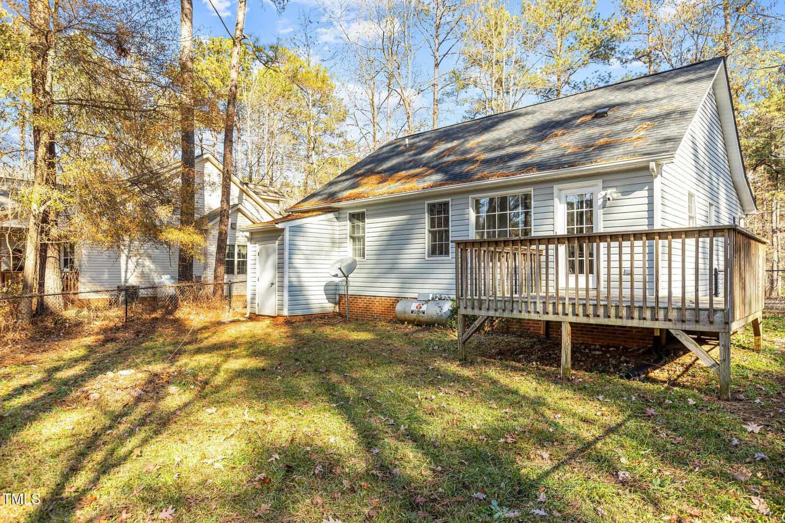 Small white house with a wooden deck in a grassy, leaf-covered backyard surrounded by tall trees on a sunny day.