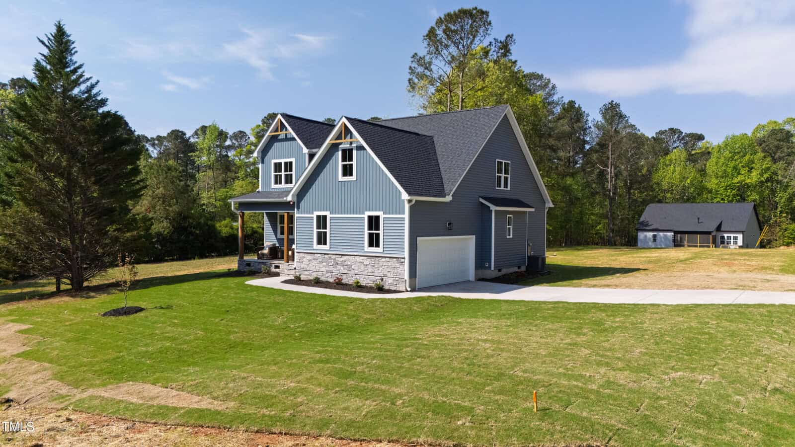 A modern blue-gray two-story house with a front porch, attached garage, and neatly landscaped lawn sits in a spacious yard with trees and a smaller building in the background under a blue sky.