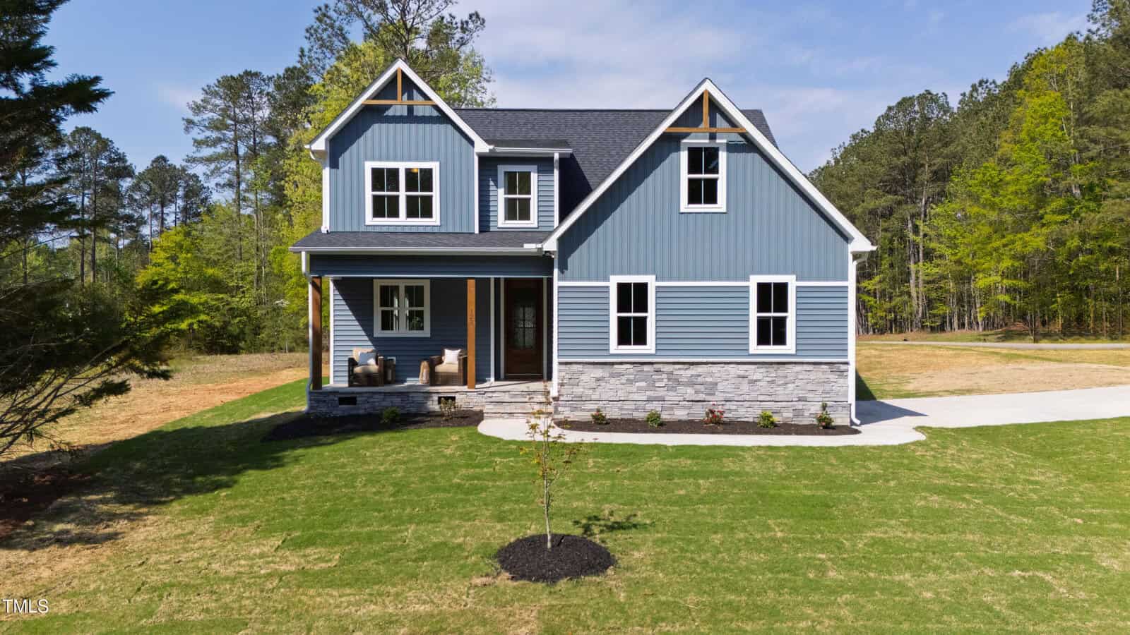A two-story blue house with white trim, a covered front porch, stone accents, and a neatly manicured lawn awaits you at 123 Wichita Way in Louisburg. This charming home is surrounded by trees under a clear blue sky.