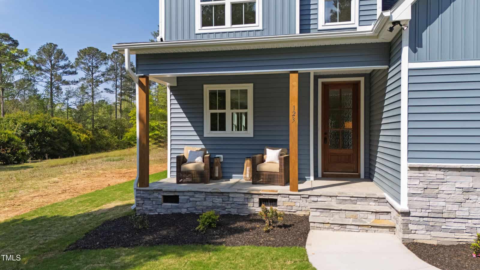 A modern house with blue siding, stone accents, a wooden front door, and a covered porch featuring two wicker chairs with cushions. The lawn is partially landscaped with shrubs and trees in the background.