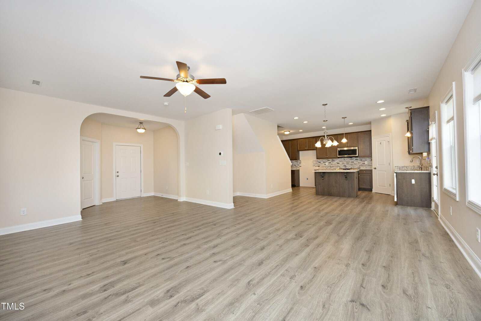 A spacious, open-concept living area with light wood flooring. It features a ceiling fan, recessed lighting, and a view into a modern kitchen with an island and pendant lights. White walls and large windows provide natural light.