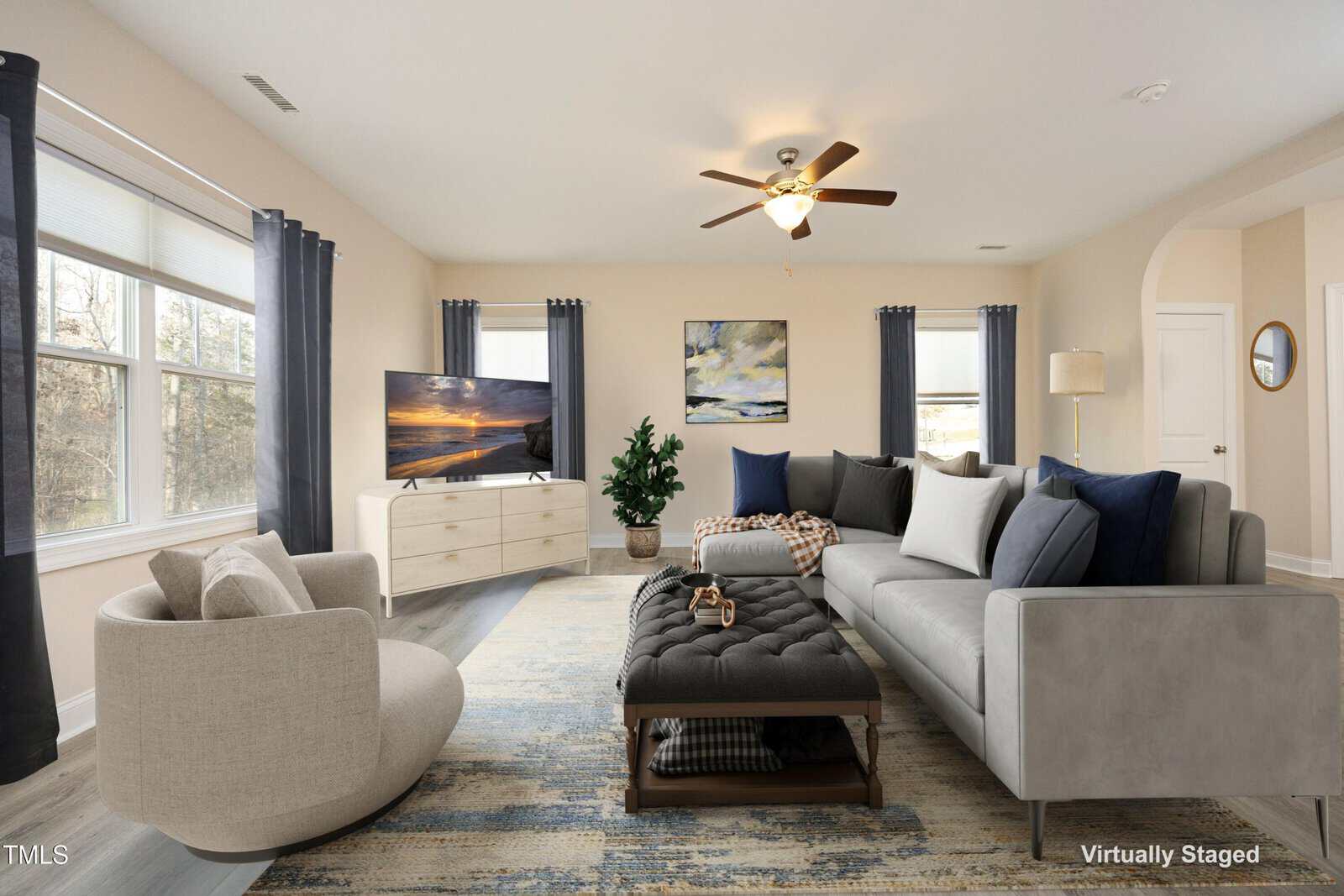 A virtually staged living room with beige walls, large windows, and a ceiling fan. The room features a gray sofa, a round armchair, a tufted ottoman, a TV on a stand, and a large rug. A potted plant and wall art add decorative touches.