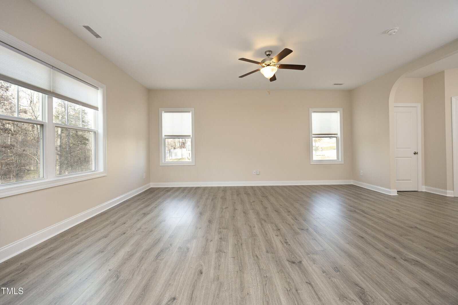 Spacious empty room with light wood flooring and beige walls. Two large windows bring in natural light. A ceiling fan is centered on the ceiling. Doorways lead to adjoining rooms.