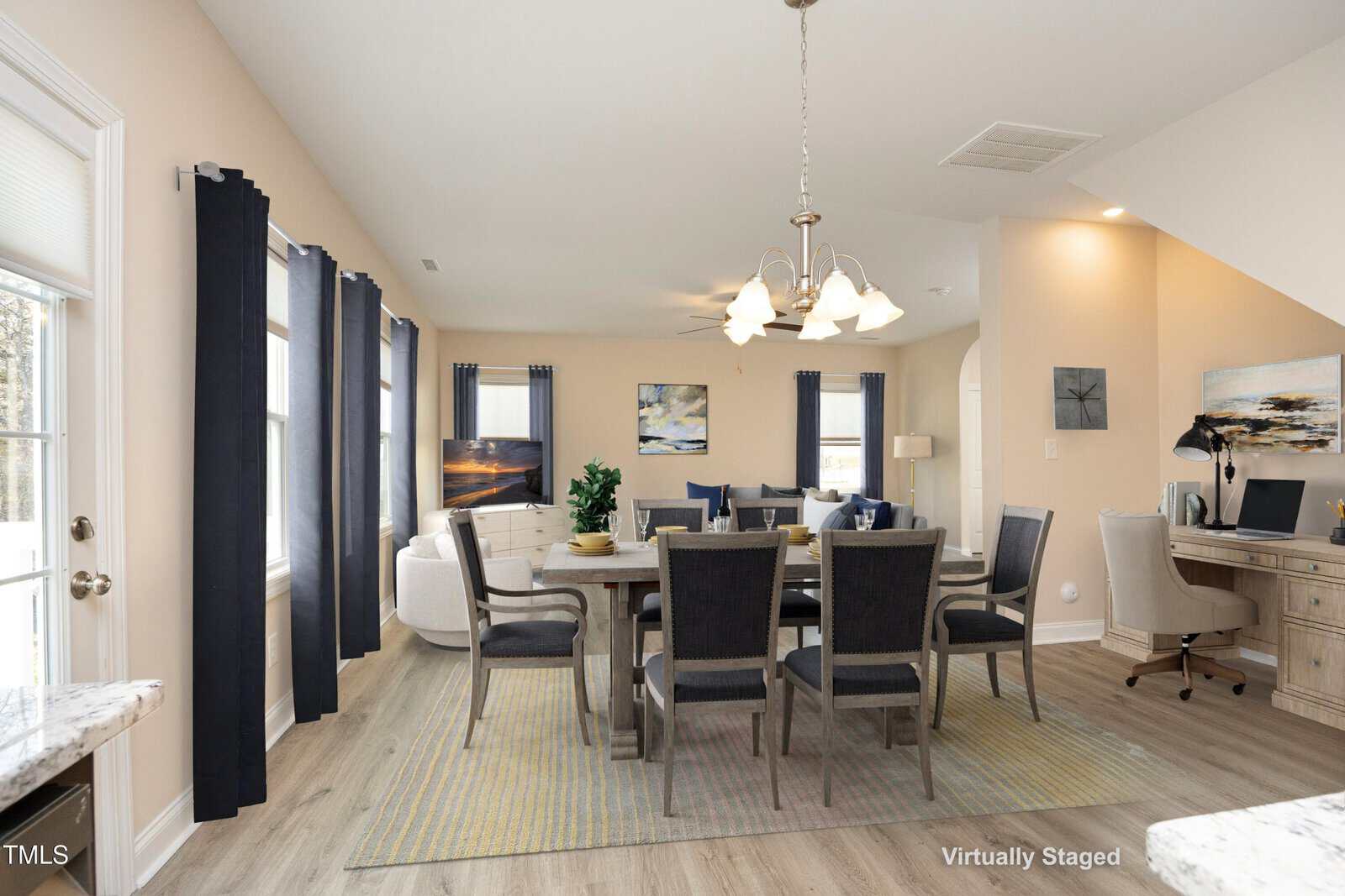 A virtually staged dining room with a wooden table and eight chairs. A modern chandelier hangs above, and a large window with dark curtains lets in natural light. Wall art and a small desk with a computer are visible in the background.