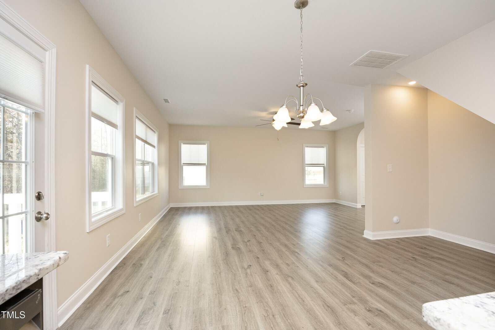 Spacious, empty room with light wood flooring, beige walls, and several windows with white blinds. A ceiling chandelier is visible, with a partial view of a staircase on the right.