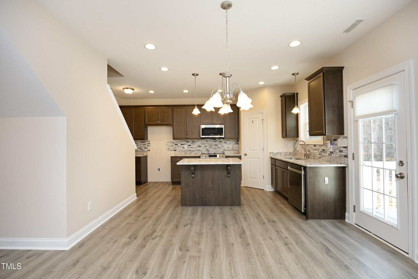 Modern kitchen with wood cabinets, island, and stainless steel appliances. Light wood flooring, pendant lighting, and large windowed door to the right. Neutral wall color and white trim throughout the room.