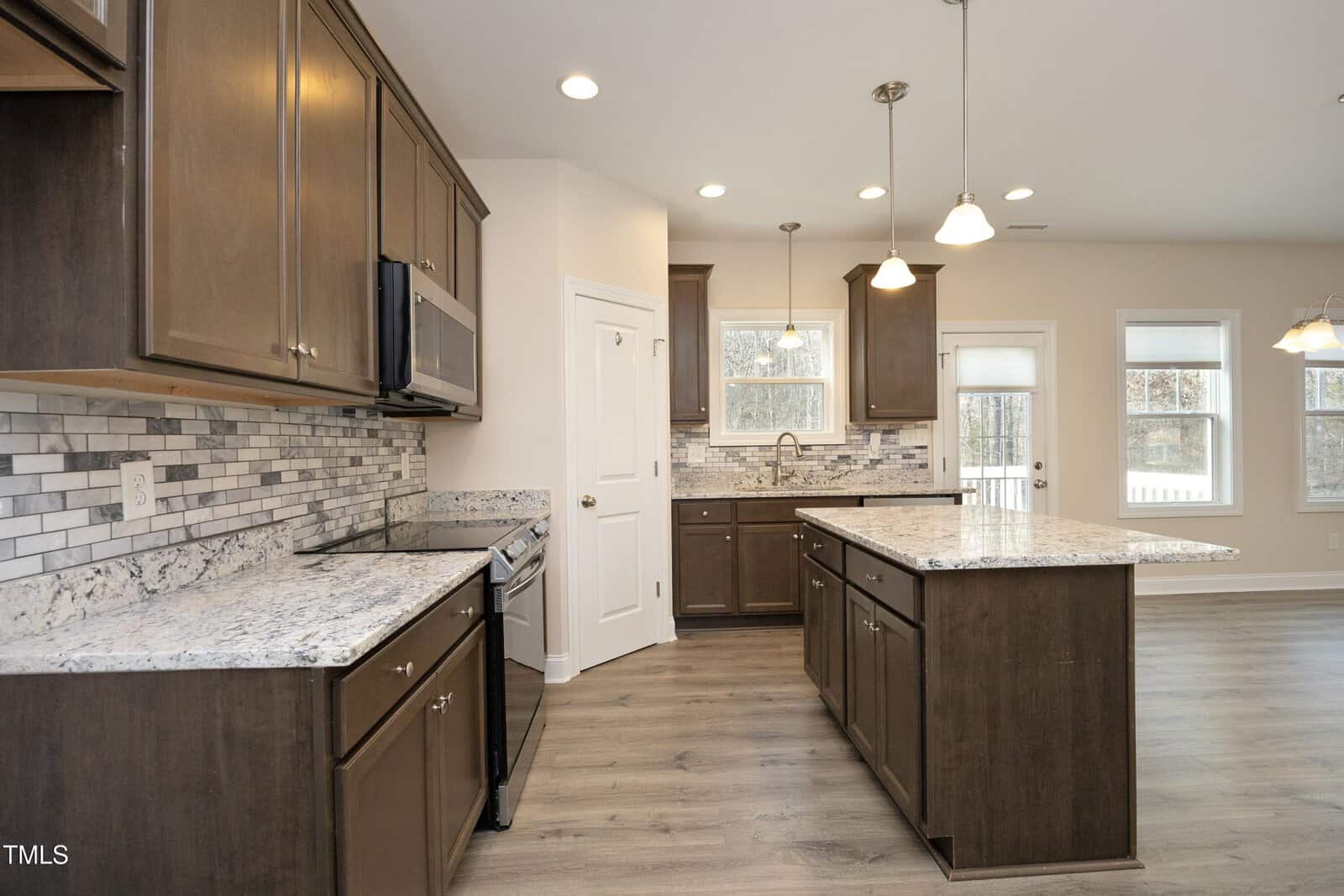 Modern kitchen with dark wood cabinets, granite countertops, stainless steel appliances, tiled backsplash, and pendant lights over a large island. Two windows above the sink and large windows in the adjacent dining area provide natural light.
