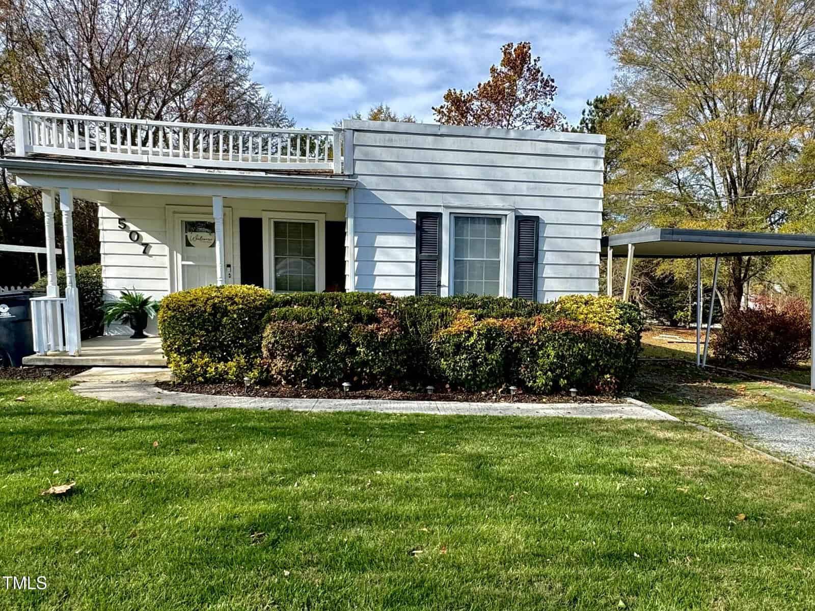 A small, single-story white house at 507 N Mack Street in Creedmoor features a flat roof and rooftop deck. It boasts a front porch, dark shutters, and is surrounded by a neatly trimmed hedge. The grassy yard includes a carport on the right side, under a partly cloudy sky.