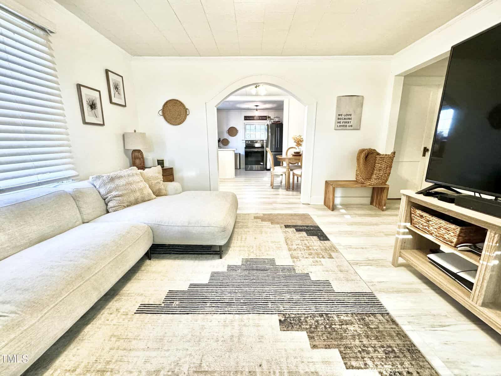 A cozy living room with a light-colored sectional sofa on a patterned rug. There is a large flat-screen TV on a wooden stand. The space opens to a kitchen through an arched doorway. White walls and light wood floors enhance the bright, airy feel.
