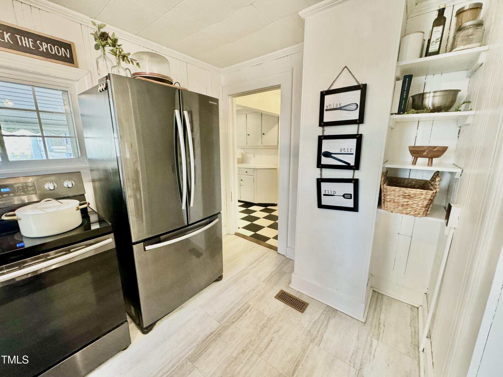 A kitchen with a stainless steel refrigerator and stove. A pot sits on the stovetop. Wall art with kitchen utensils hangs next to built-in shelves holding various items. The floor leads to another room with checkered flooring.