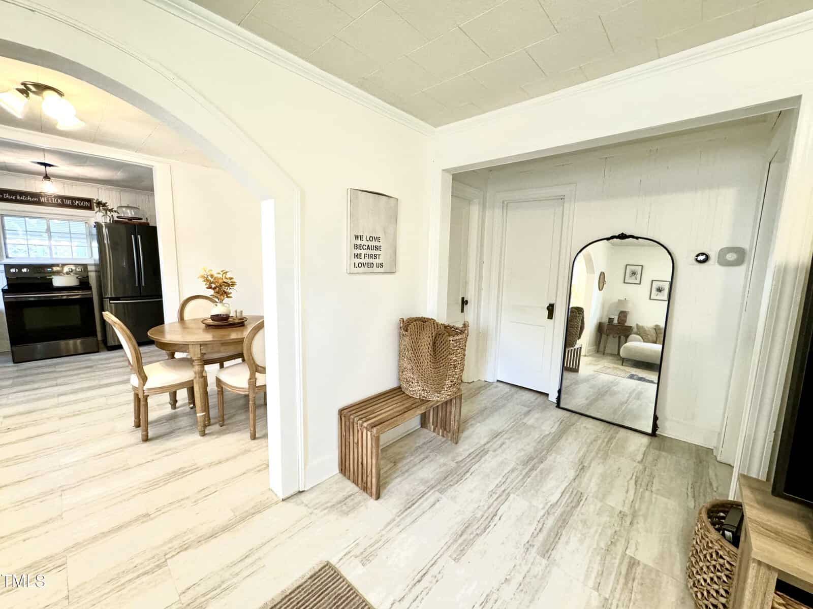 A bright room with light-colored flooring and walls. A wooden bench and two wicker baskets are near a large mirror. Adjacent is a dining area with a round table and chairs, leading to a kitchen with dark appliances. A sign on the wall reads about love.