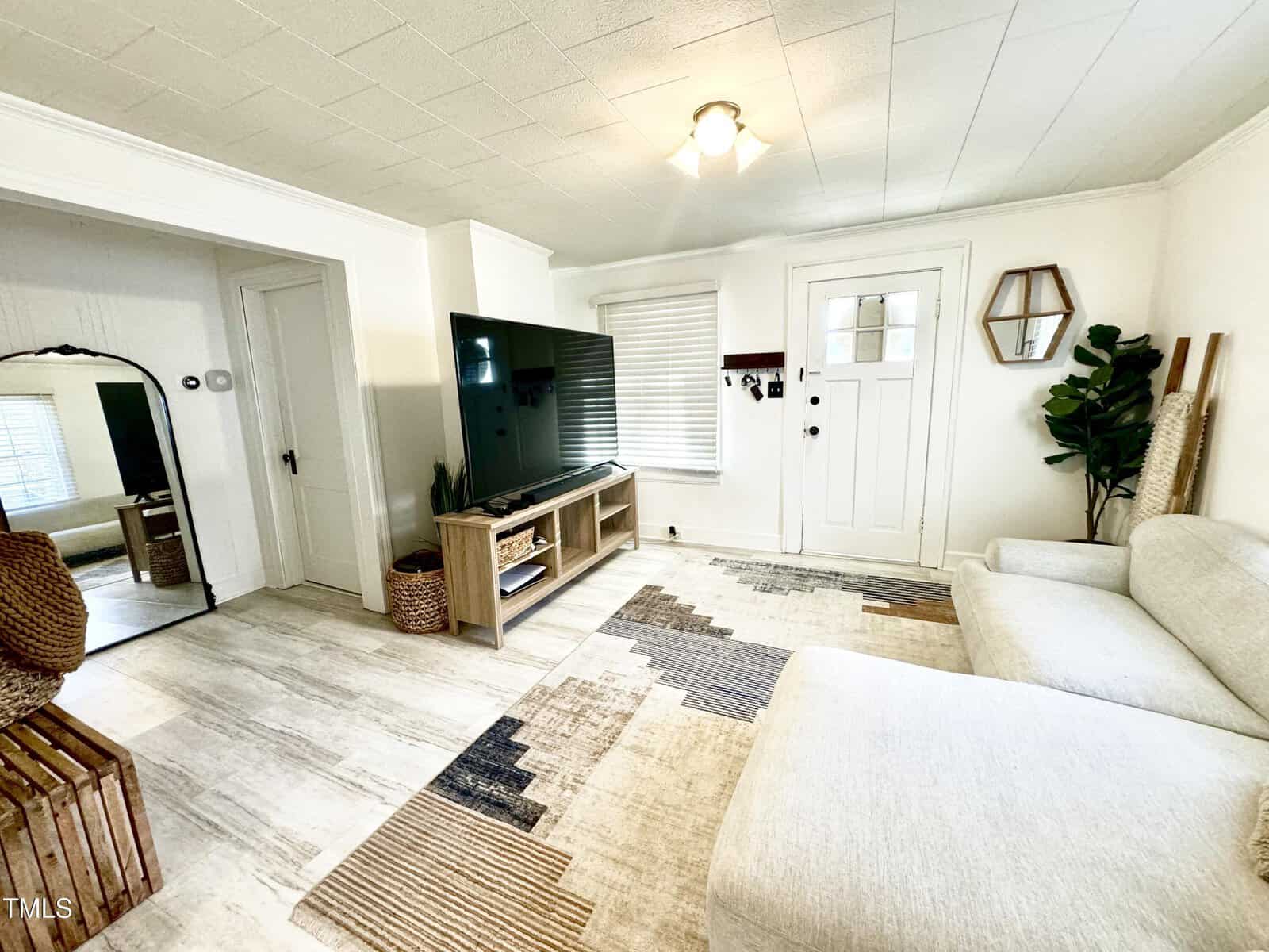 A minimalist living room features a light-colored sectional sofa, a flat-screen TV on a wooden stand, and a geometric rug. The room has white walls, a window with blinds, a mirror, and a potted plant in the corner.