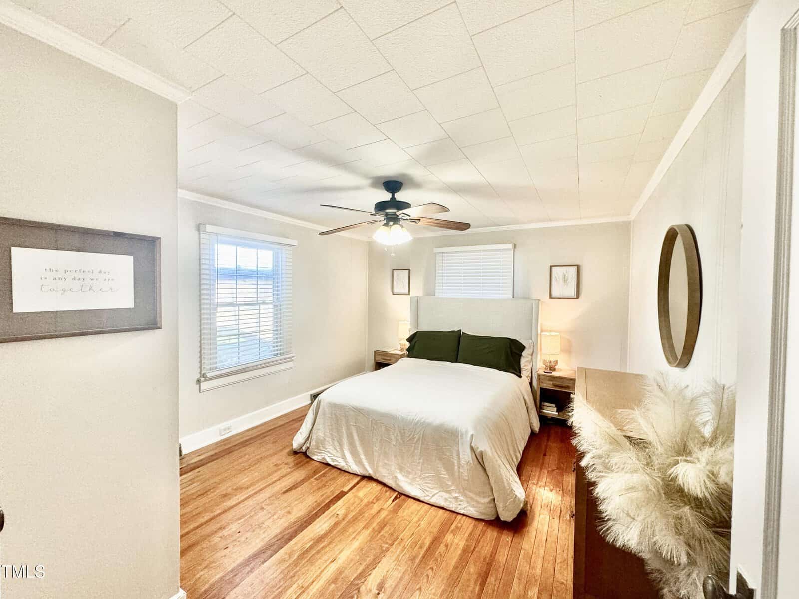 A cozy bedroom features a neatly made bed with white linens and dark cushions. A ceiling fan with lights is centered above. Wall art and a window with blinds adorn the walls. A bunch of decorative pampas grass stands in the corner.