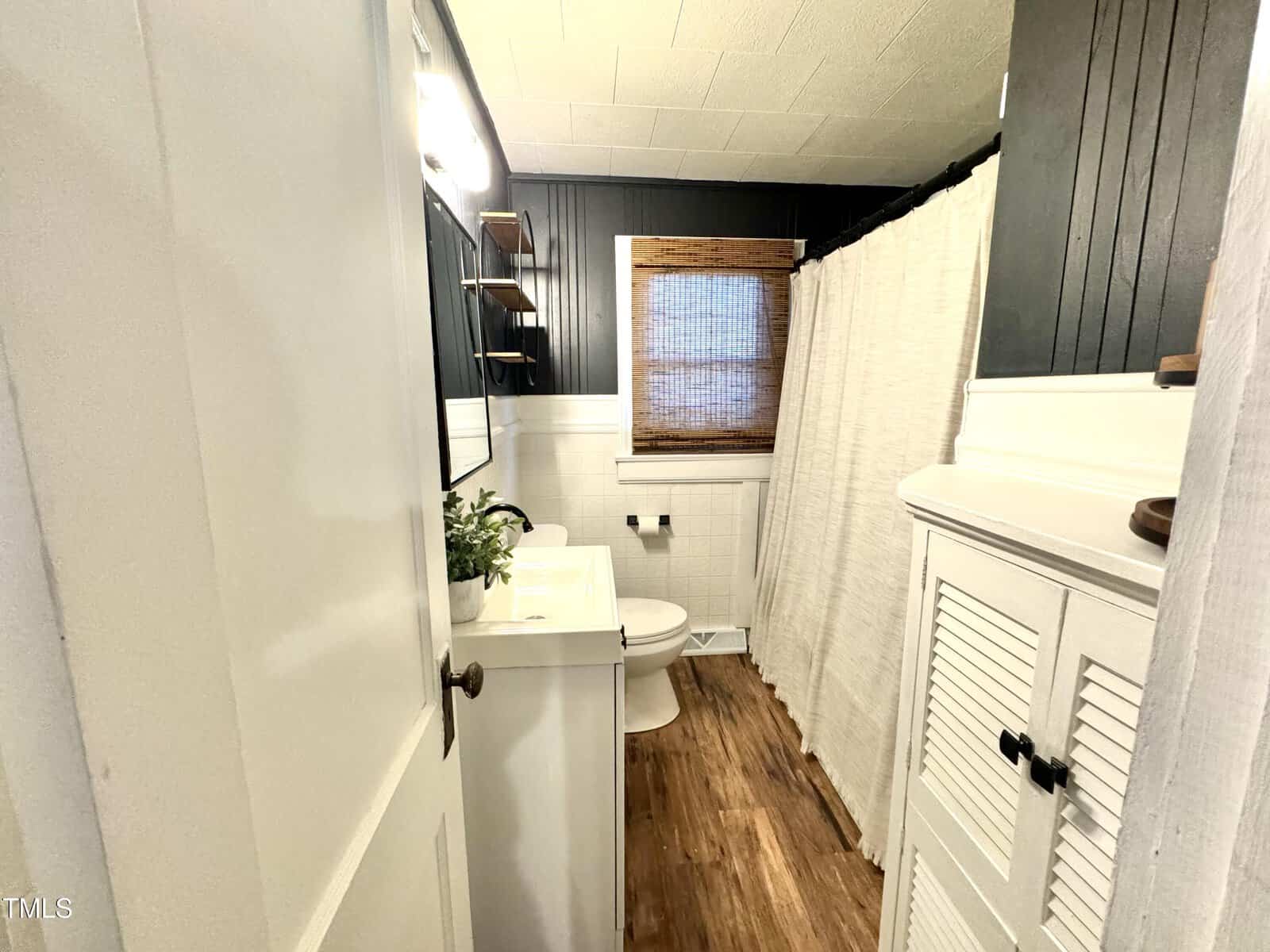 A small bathroom with wood flooring, white walls, and dark wall paneling. It features a white vanity with a mirror, a toilet, a shower with a beige curtain, a window with a bamboo shade, and a white cabinet with louvered doors.